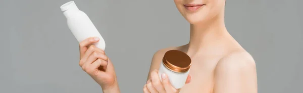 Panoramic shot of cheerful woman holding container with face cream and bottle with body lotion isolated on grey — Stock Photo