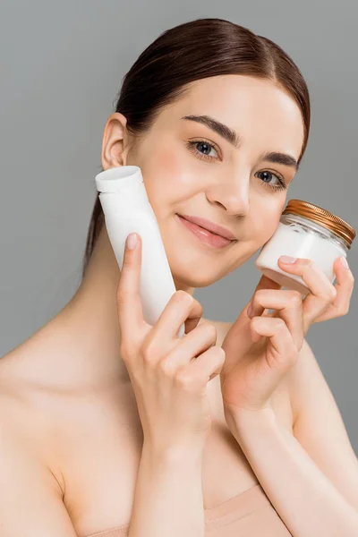 Cheerful woman holding container with face cream and bottle with body lotion isolated on grey — Stock Photo