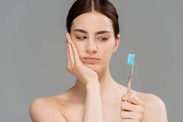 Молода гола жінка торкається обличчя і дивиться на зубну щітку ізольовано на сірому — стокове фото