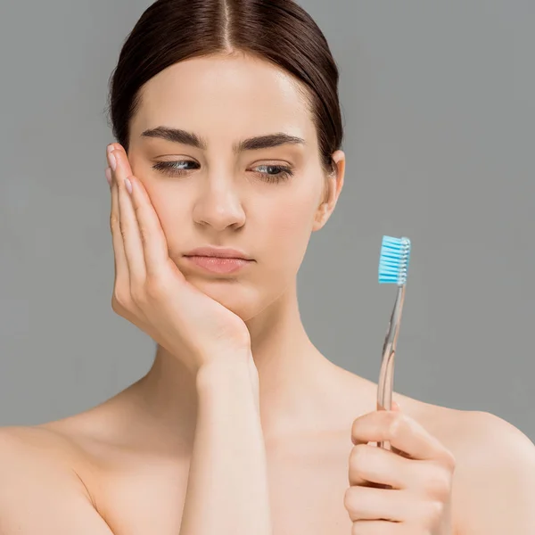 Attractive naked woman touching face and looking at toothbrush isolated on grey — Stock Photo