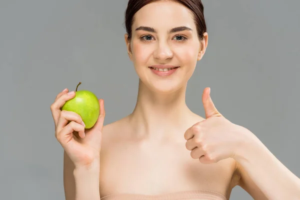 Glückliche Frau lächelt, während sie grünen Apfel in der Hand hält und den Daumen vereinzelt auf grau zeigt — Stockfoto