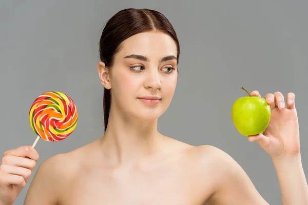 Atractiva chica desnuda mirando a la manzana verde mientras sostiene dulce piruleta aislado en gris - foto de stock