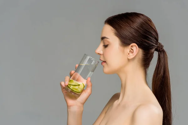Vue latérale d'une jolie femme nue regardant un verre d'eau avec de la chaux tranchée isolée sur du gris — Photo de stock