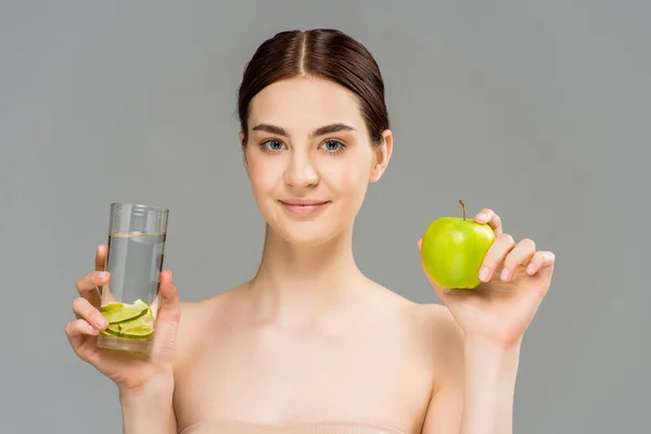Cheerful girl holding glass of water with sliced lime and green apple isolated on grey — Stock Photo