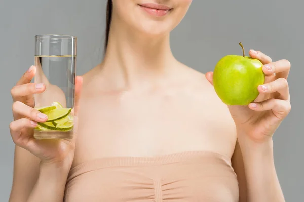 Vista recortada de niña alegre sosteniendo vaso de agua con lima en rodajas y manzana verde aislada en gris - foto de stock
