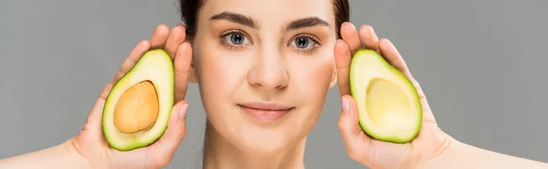 Panoramic shot of young woman holding avocado halves isolated on grey — Stock Photo