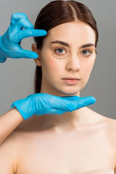 Vista recortada de cirujano plástico en guantes de látex cerca de mujer atractiva con marcas en la cara aislada en gris - foto de stock