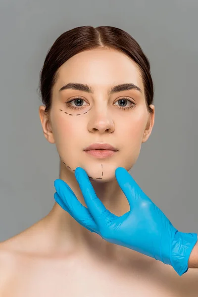 Обрезанный вид пластического хирурга в латексной перчатке трогательное лицо привлекательной обнаженной женщины, изолированной на серой — стоковое фото