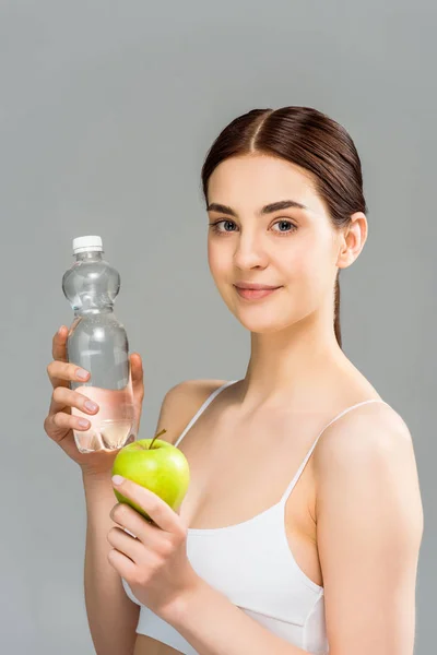 Cheerful brunette girl holding bottle with water and green apple isolated on grey — Stock Photo