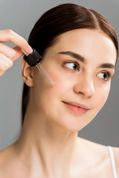 Cheerful brunette woman holding pipette while applying serum on face isolated on grey — Stock Photo