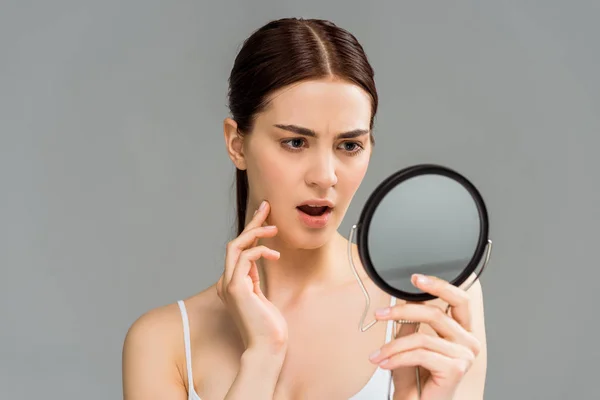 Потрясенная молодая женщина, смотрящая на зеркало, изолированное от серого — стоковое фото
