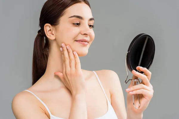 Jolie jeune femme regardant miroir et visage touchant isolé sur gris — Photo de stock