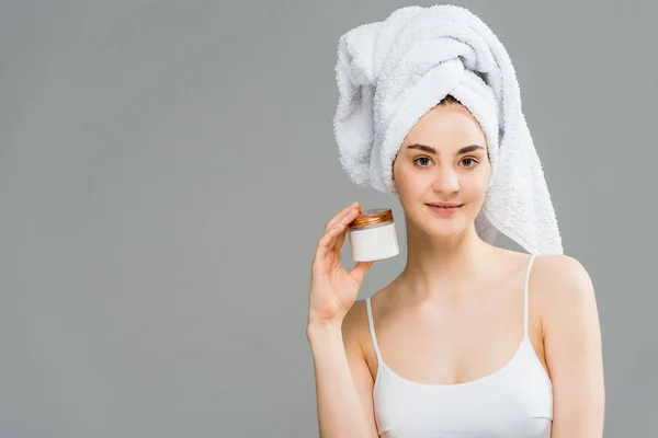 Mujer alegre con toalla en la cabeza sosteniendo contenedor con crema cosmética aislado en gris - foto de stock