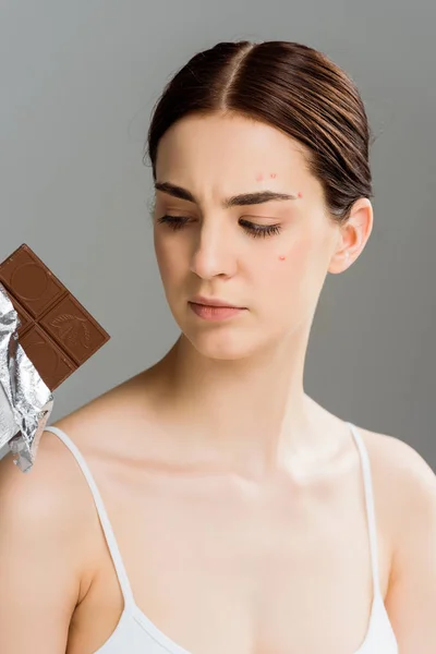 Bouleversé jeune femme brune avec de l'acné sur le visage en regardant la barre de chocolat isolé sur gris — Photo de stock