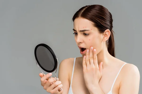 Shocked young woman with acne on face looking at mirror isolated on grey — Stock Photo