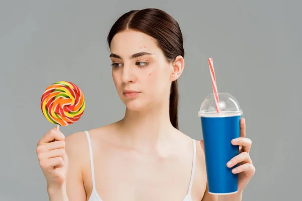Young brunette woman with acne on face holding sweet lollipop and plastic cup isolated on grey — Stock Photo