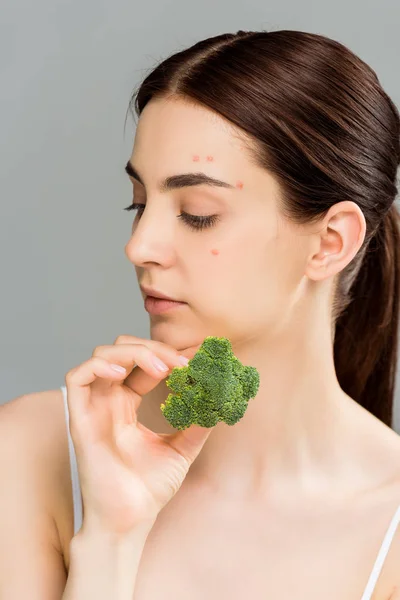 Young brunette woman with acne on face holding green broccoli isolated on grey — Stock Photo