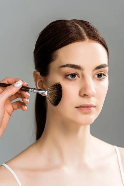 Cropped view of makeup artist holding cosmetic brush near cheek of attractive woman isolated on grey — Stock Photo