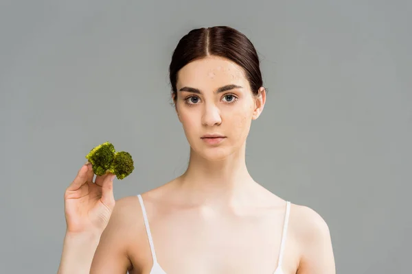 Brunette woman with face scrub holding green broccoli isolated on grey — Stock Photo