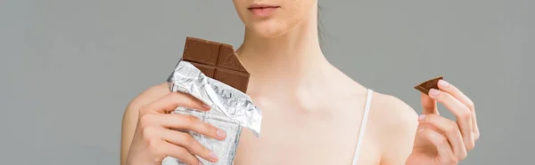 Panoramic shot of young woman with problem skin holding chocolate bar and looking at camera isolated on grey — Stock Photo