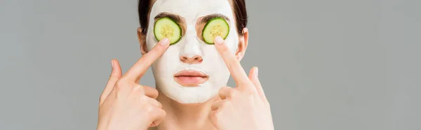 Panoramic shot of young woman with clay mask and cucumbers on face isolated on grey — Stock Photo