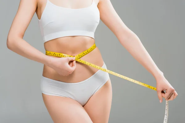 Cropped view of young woman in underwear standing and measuring body with measuring tape isolated on grey — Stock Photo