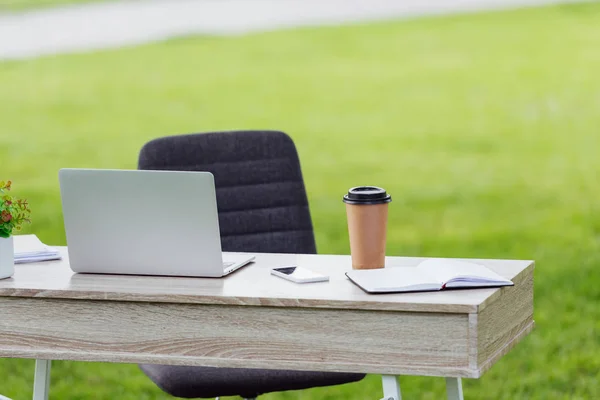 Офісний стіл з ноутбуком, кавою, щоб піти, блокнот і смартфон біля офісного крісла в парку — стокове фото