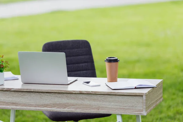 Office table with laptop, coffee to go, notebook and smartphone near office chair in park — Stock Photo