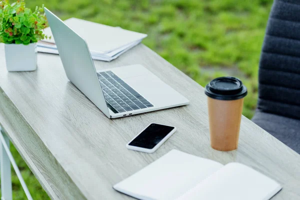Selective focus of office table with laptop, coffee to go, notebook and smartphone near office chair in park — Stock Photo