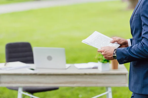 Ausgeschnittener Blick auf jungen Geschäftsmann, der Zeitung hält und im Park am Tisch steht — Stockfoto