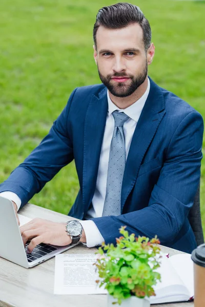 Homem de negócios bonito usando laptop, sentado no parque atrás da mesa e olhando para a câmera — Fotografia de Stock