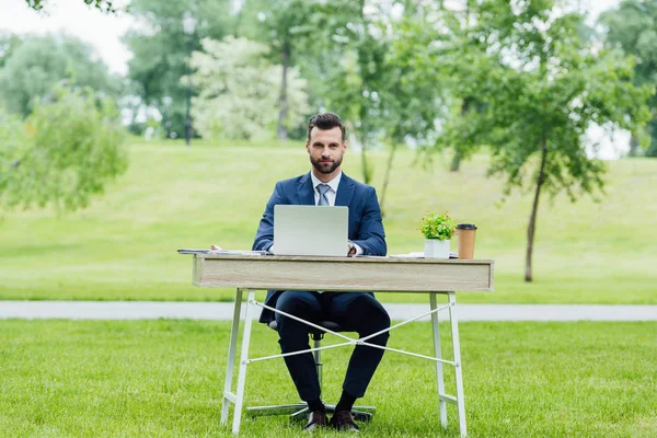 Handsome young businessman in formal wear using laptop, sitting at table in park and looking at camera — Stock Photo