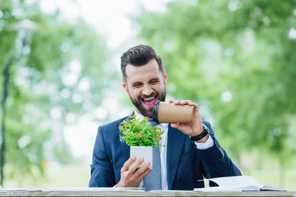 Sonriente joven empresario guapo sosteniendo café para ir y plantar - foto de stock