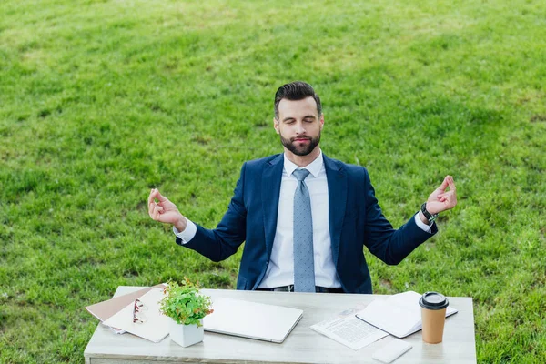 Schöner junger Geschäftsmann meditiert, während er im Park hinter einem Tisch sitzt — Stockfoto