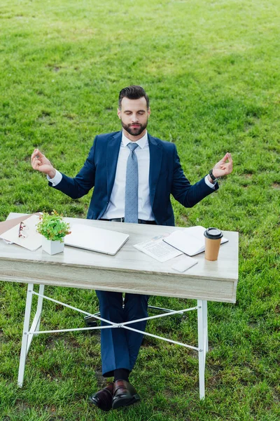 Full length view of handsome young businessman in formal wear meditating while sitting at table with various office stuff in park — Stock Photo