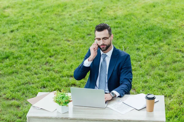 Joven hombre de negocios en ropa formal usando el ordenador portátil y hablando en el teléfono inteligente mientras está sentado en el parque detrás de la mesa de la oficina - foto de stock