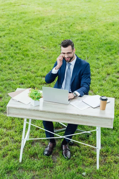 Joven hombre de negocios en ropa formal usando el ordenador portátil, sonriendo y hablando en el teléfono inteligente mientras está sentado en el parque detrás de la mesa de la oficina — Stock Photo