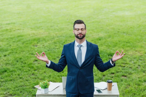 Young businessman in formal wear meditating near table with various office stuff in park — Stock Photo