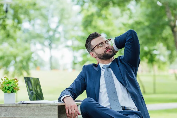 Young businessman in formal wear sitting at table with laptop, plant and notebook and relaxing in park — Stock Photo