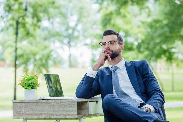 Joven hombre de negocios sentado en la mesa con el ordenador portátil y la planta en el parque y mirando hacia otro lado - foto de stock