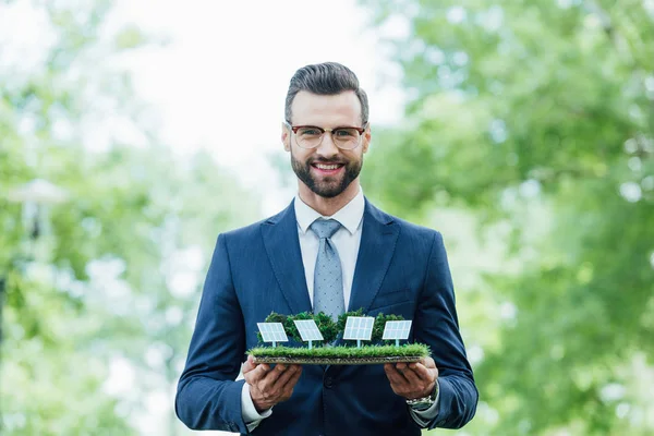 Молодой бизнесмен, держащий макет парка с солнечными батареями, улыбающийся и смотрящий в камеру, стоя в парке — стоковое фото