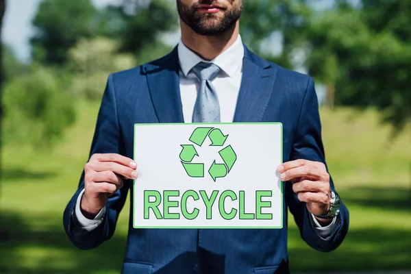 Cropped view of young businessman holding card with recycle inscription while standing in park — Stock Photo