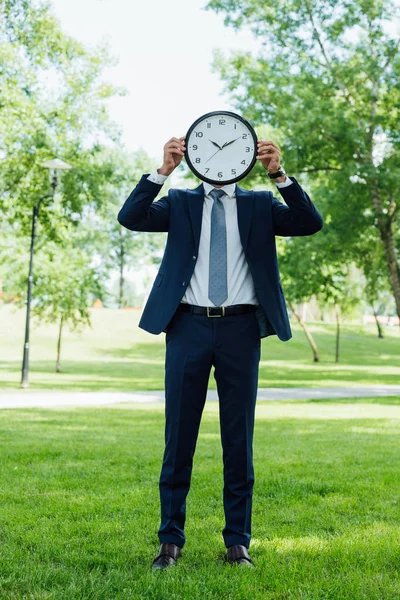 Vue pleine longueur de jeune homme d'affaires couvrant visage avec horloge dans le parc — Photo de stock
