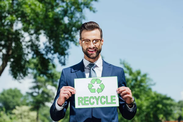 Junger Geschäftsmann hält Karte mit Recycling-Aufschrift, blickt in die Kamera und lächelt — Stockfoto