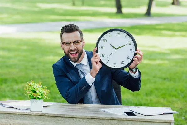 Businessman sitting behind table with flowerpot, smartphone and notebooks, holding clock and looking at camera — Stock Photo