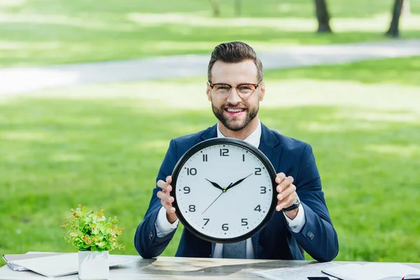 Hombre de negocios sentado detrás de la mesa con la planta y los cuadernos, sonriendo, mirando a la cámara y sosteniendo el reloj - foto de stock