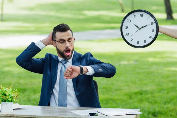 Sorprendido joven hombre de negocios sentado en la mesa con la planta, teléfono inteligente y portátiles, sosteniendo la cabeza y mirando el reloj - foto de stock