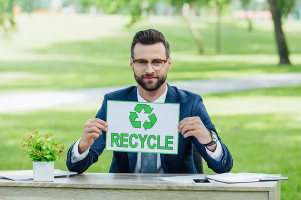 Young businessman sitting at table with plant, smartphone and notebooks in park, holding card with recycle inscription and looking at camera — Stock Photo