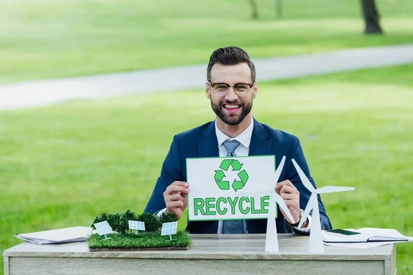 Jeune homme d'affaires à table avec des moulins à vent et des batteries solaires mises en page tenant carte avec inscription de recyclage — Photo de stock