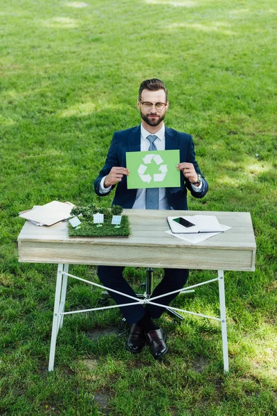 Winkelaufnahme eines jungen Geschäftsmannes, der am Tisch mit Windmühlen sitzt, Karte mit Recyclingschild in der Hand hält und in die Kamera blickt — Stockfoto